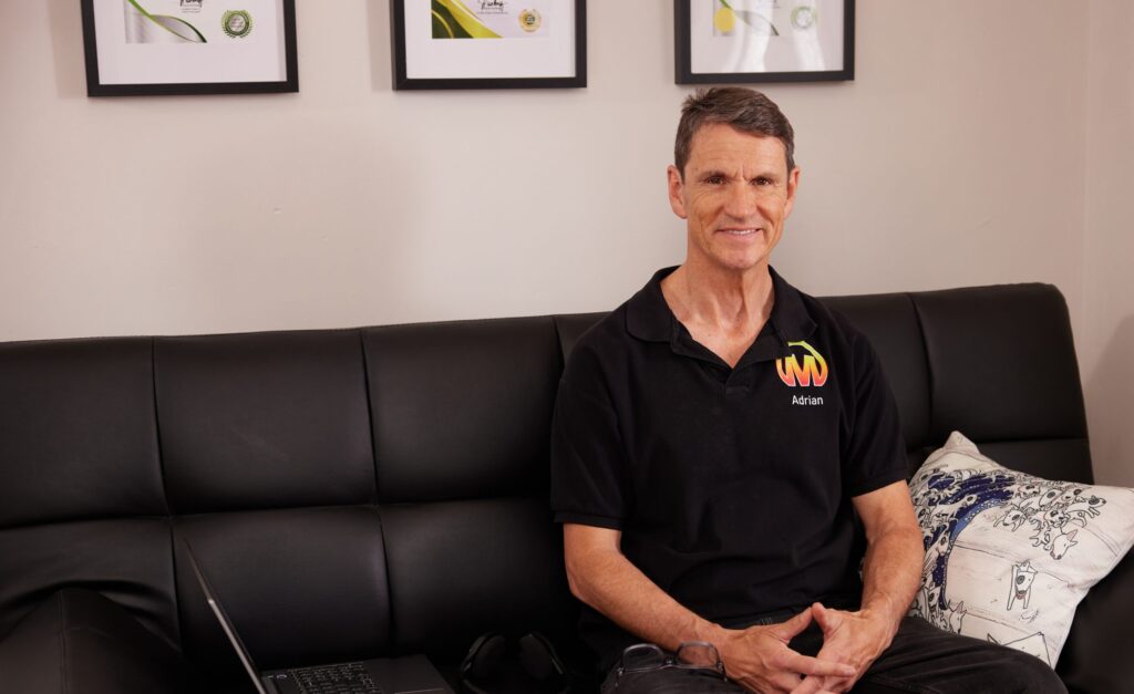 Contact Adrian Spear who is sitting on a black leather lounge with certificates hanging on the wall.