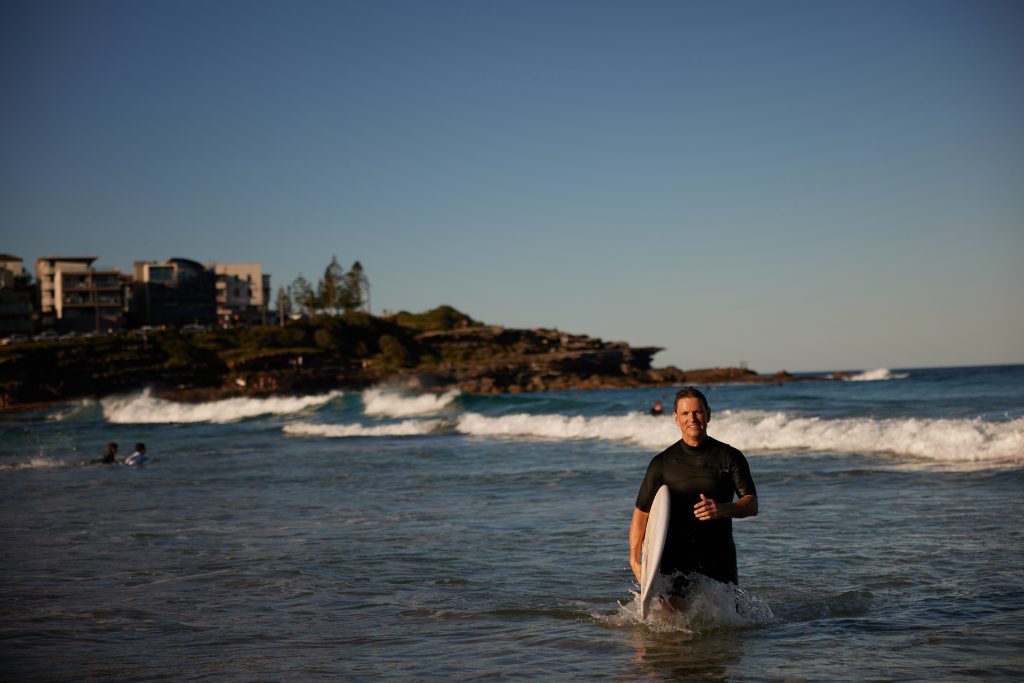 Adrian Spear with a surfboard under his arm walking out of the surf.