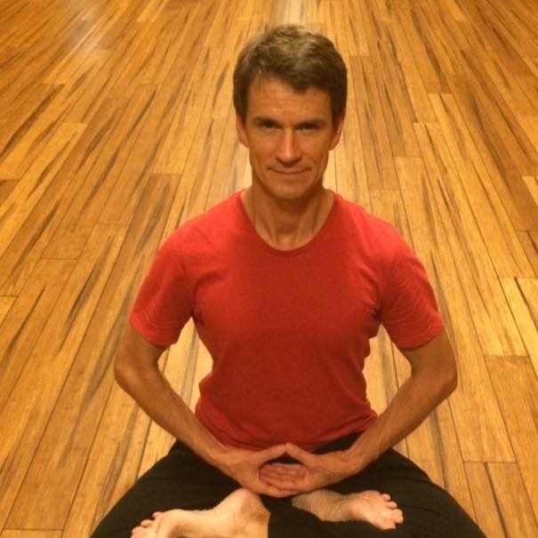 mindfulness meditation expert Adrian Spear meditatiing on a wooden floor wearing a red shirt and black pants 